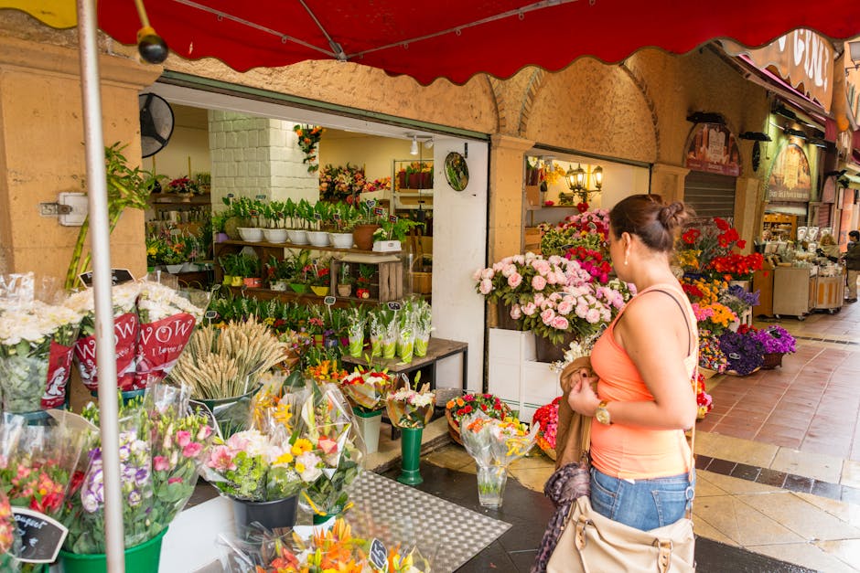 A woman in an orange sleeveless top and blue jeans is standing outside a flower shop, holding a small bouquet of mixed flowers. In front of her, there are various floral arrangements and bouquets wrap