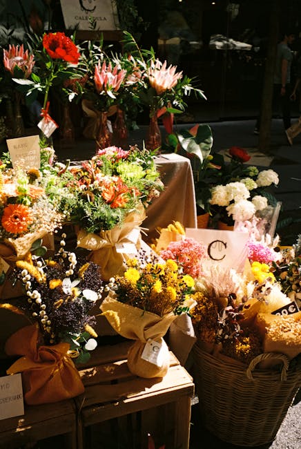 An arrangement of various fresh floral bouquets displayed on a wooden table outdoors, featuring a mix of blossoms including pink lilies, white roses, yellow daisies, orange gerbera daisies, and sprigs